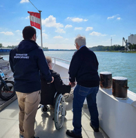 Zwei Menschen helfen einer Person im Rollstuhl auf einem Boot.