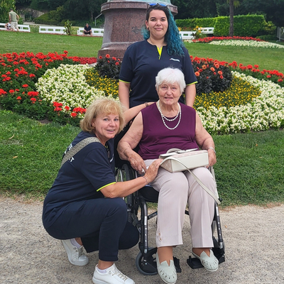 Drei Frauen, eine im Rollstuhl, posieren lächelnd vor bunten Blumenbeeten.
