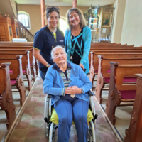 Drei Frauen, eine im Rollstuhl, stehen in einer Kirche zusammen.