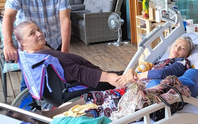 Eine Frau im Rollstuhl hält die Hand einer liegenden Frau, während ein Mann lächelt.