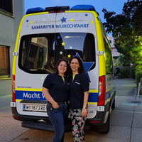 Zwei Frauen stehen lächelnd vor einem Krankenwagen mit dem Schriftzug „Samariter-Wunschfahrt“.