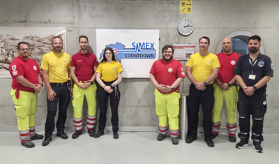 Gruppenfoto vor dem Simexschild Gruppenbild von Rettungskräften mit unterschiedlichfarbigen Uniformen vor einem Countdown-Schild.