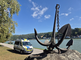 Ein großer Anker steht am Ufer eines ruhigen Gewässers mit einem Fahrzeug im Hintergrund.
