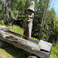 Holzskulptur eines Mannes mit Hut, der aus einer Holzrinne Wasser trinkt.
